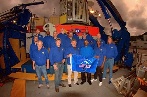The 2018 discovery team on deck at dusk in front of the launch system, holding the Lost 52 Project flag